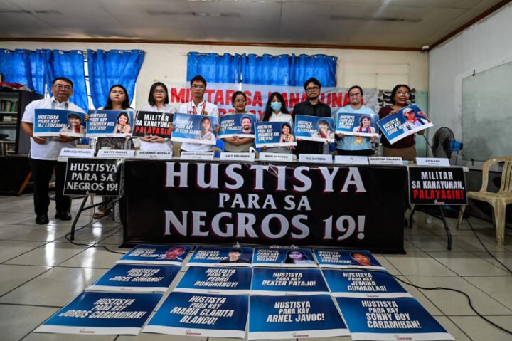 Photo of 9 people holding signs of victims of the Toboso 19 massacre. They are standing behind a black banner with white text stating "Justice for the Negros 19!" On the floor are signs indicating various calls of the organizations at the press conference.
